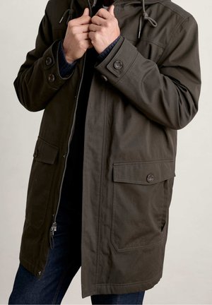 Osoba zapinająca ciemnozieloną kurtkę z kapturem, z kieszeniami na klapki zapinanymi na guziki i zamkiem błyskawicznym, ubrana w ciemnoniebieskie dżinsy i granatowy sweter.