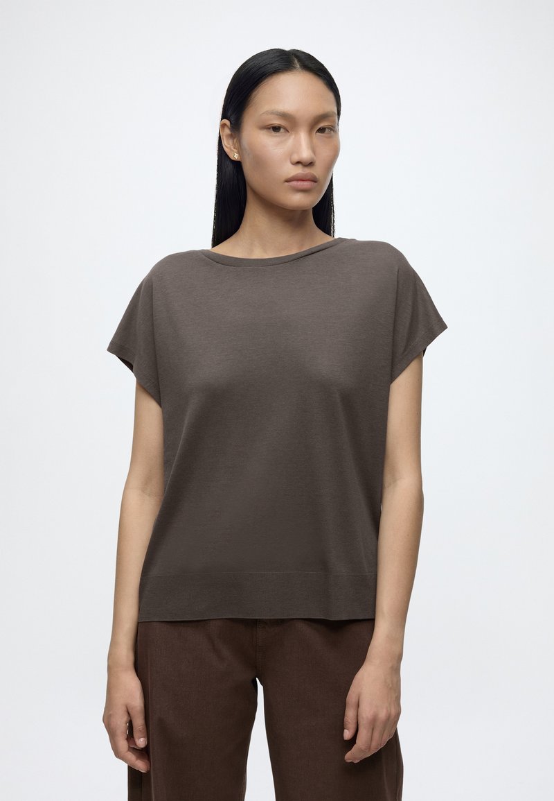 Femme aux cheveux noirs et raides, portant un haut marron ample à manches courtes et un pantalon marron, debout devant un fond blanc uni.