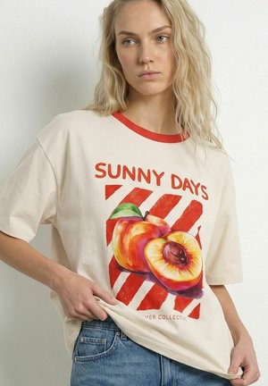 Kvinde med langt blond hår iført en cremefarvet t-shirt med teksten "Sunny Days" og en ferskengrafik, kombineret med blå jeans mod en ensfarvet baggrund.