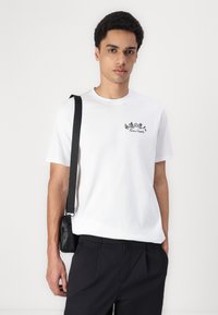 Hvid t-shirt med korte ærmer og sorte grafikker, iført med sorte bukser. Har en lille skuldertaske med en rem. Minimalistisk design.