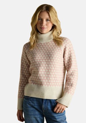 Young woman with blonde hair wearing a pink and cream patterned turtleneck sweater and blue jeans, standing against a plain background.