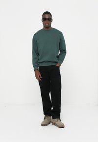 Donkergroen sweatshirt, ronde halslijn, ribgebreide manchetten, zwart logo-embleem; zwarte broek, lichtgrijze laarzen met veters, gedragen met zonnebril.