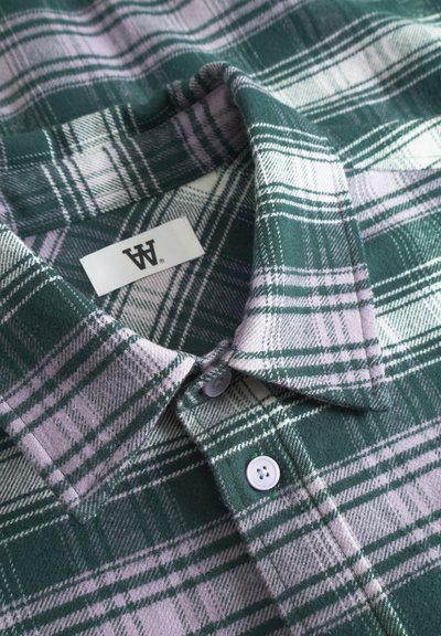 Chemise en flanelle à carreaux verts et lilas avec un col, dotée de boutons blancs et d'une étiquette. Texture douce et motif à carreaux.