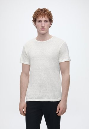 Jeune homme aux cheveux roux bouclés portant un t-shirt blanc uni à manches courtes et un pantalon noir, se tenant devant un fond blanc.