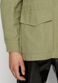 Olive green canvas jacket with two front pockets, fitted cuffs, and visible stitching details. Paired with shiny black pants.