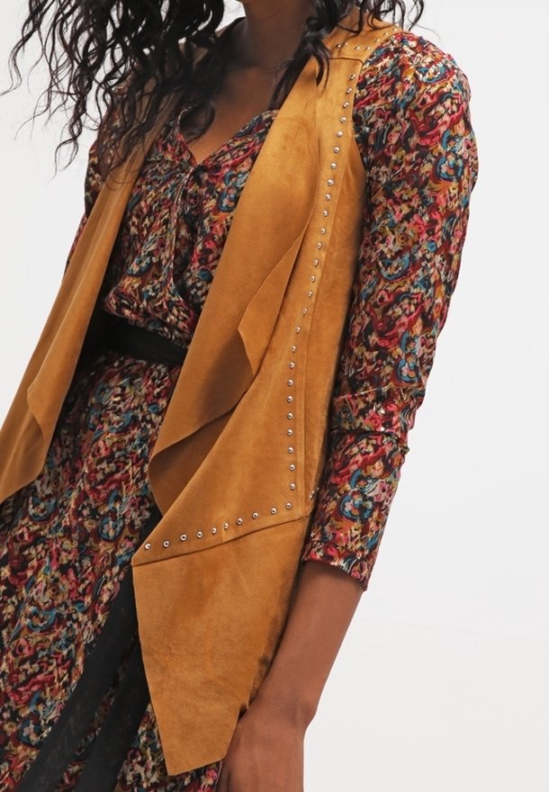 Píseňová bezrukávová vesta z broušené kůže s volným designem, zdobená stříbrnými cvoky podél okrajů, nošená přes barevné šaty s květinovým potiskem.