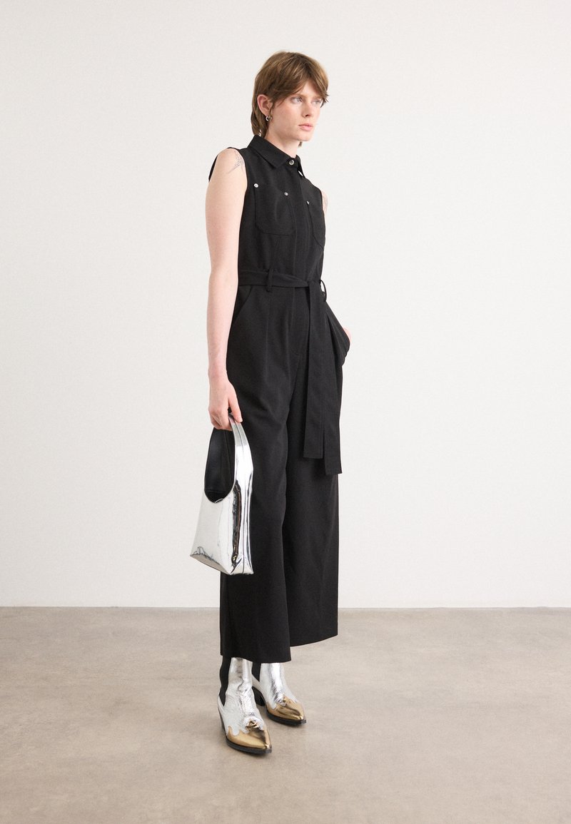 Mono sin mangas negro con bolsillos frontales y cintura anudada, bolso de mano metálico plateado y botas cortas metálicas brillantes con acentos dorados.