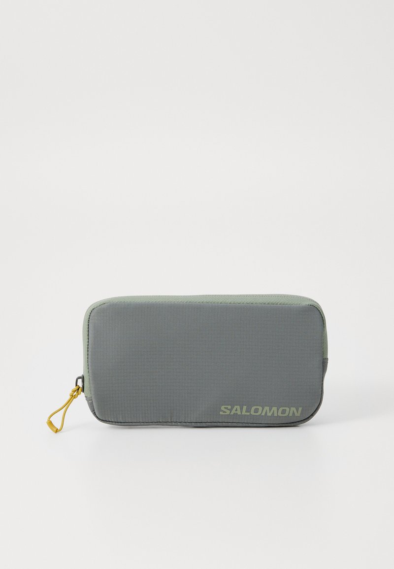 Šedý obdĺžnikový vankúšik vyrobený z textúrovaného nylonu s žltým zipsom. Na prednej strane sa nachádza logo "SALOMON" v svetlozelenej farbe.