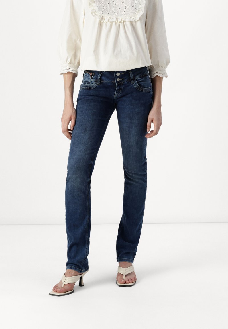 Vaqueros azul oscuro con un diseño ajustado, cinco bolsillos y costuras en contraste. Llevado con una blusa blanca ligera y texturizada, y sandalias.