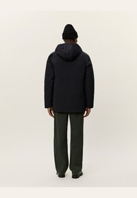 Chaqueta negra aislante con capucha, con una textura suave y corte recto. Llevada sobre pantalones verdes, destacando la vestimenta práctica para actividades al aire libre.
