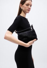 Μαύρη υφασμάτινη τσάντα με λείο, καμπυλωτό σχήμα και λεπτή λαβή, κρατημένη μπροστά από μοντέλο που φορά εφαρμοστό μαύρο φόρεμα.