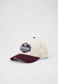 Krémová a burgundská čiapka s bočnými sieťovinami, s prominentným vyšívaným logom BRIXTON na prednej strane a zahnutým brnením.