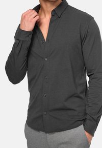 Tummanharmaa pitkähihainen napillinen paita, jossa on pehmeä tekstuuri, kapea kaulus ja mustat napit. Paidassa on istuva malli ja yksinäppäimiset kalvosimet.