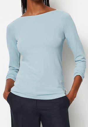 Vrouw draagt een lichtblauw, gestructureerd shirt met lange mouwen en een boothals, en een donkere broek, met handen in de zakken, staand tegen een effen achtergrond.