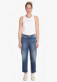 Pantalones cargo de mezclilla en azul medio con múltiples bolsillos delanteros, textura desgastada y costuras en contraste. Combinados con un top blanco sin mangas.