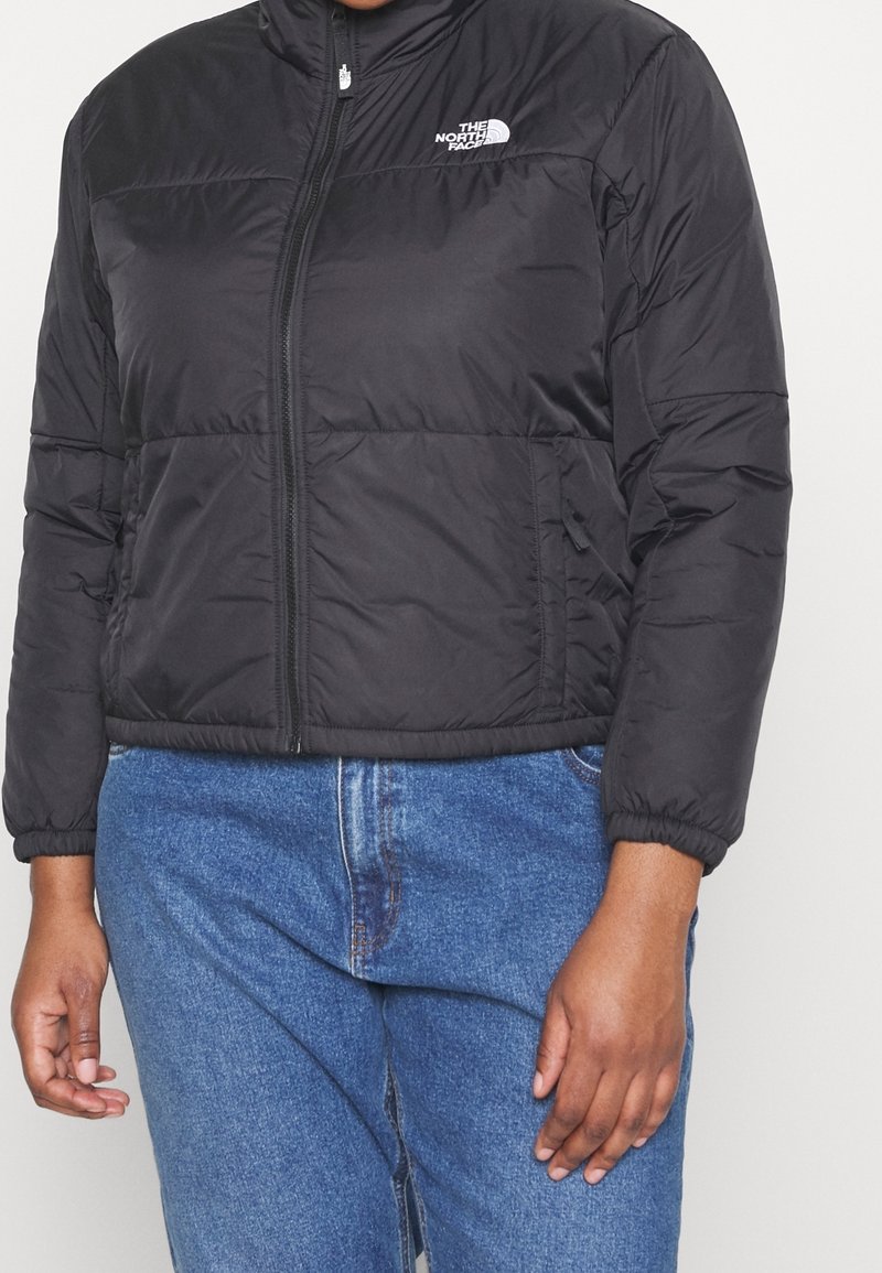 Svart pufferjacka med hög krage, dragkedja, elastiska muddar och The North Face-logotyp på vänster bröst. Buren över blå jeans.