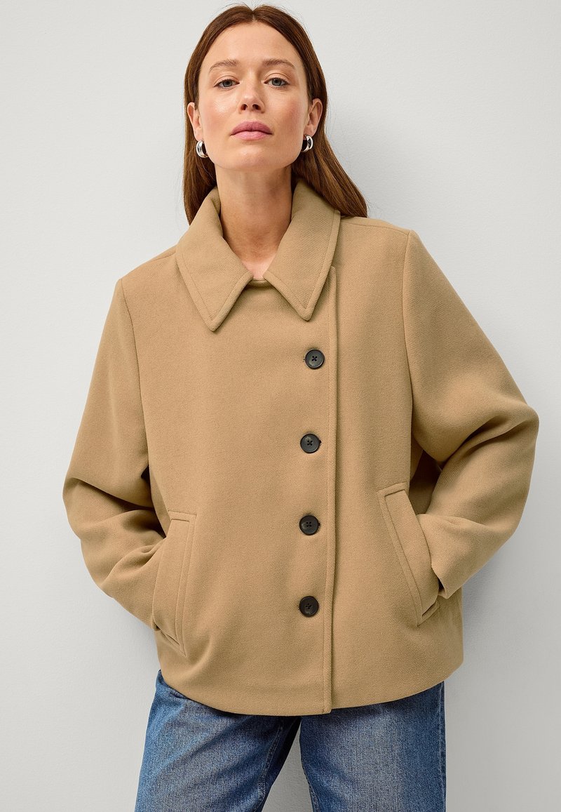 Beige uldblandet frakke med notch-krave, forlommer og fem sorte knapper, båret over blå jeans mod en neutral baggrund.