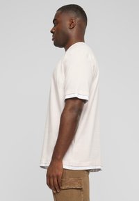 Lyserød kortærmet T-shirt med rund halsudskæring, med kontrasterende hvid kant ved ærmerne og nederste kant. Model iført brune bukser.