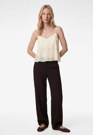 Blondynka w kremowej, bezrękawowej bluzce z szyfonu, ciemnych szerokich spodniach i brązowych balerinkach, stojąca z rękami za plecami.