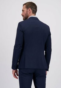 Homme aux cheveux bruns courts et à la barbe, portant un costume bleu foncé et une chemise à motifs, tourné sur le côté devant un fond uni clair.