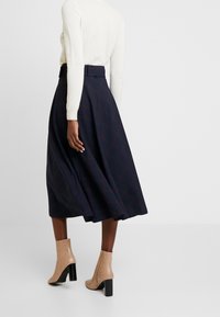 Femme portant un pull blanc, une jupe midi bleu marine à motif écossais subtil, et des bottines beiges à talons, debout devant un fond blanc.