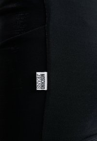 Fekete szövet, amelynek texturált felülete van, egy kiemelkedő fehér címkével, rajta a "MOSCHINO JEANS" felirat vastag betűkkel.