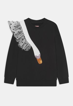 Sweat-shirt noir à manches longues avec une bretelle diagonale blanche texturée, un volant en tulle noir à pois sur l'épaule gauche et un petit cœur métallique avec une étiquette.