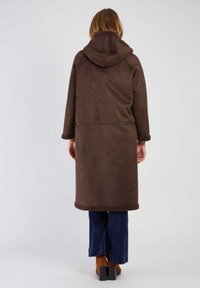 Brązowy, długi płaszcz z kapturem o miękkiej teksturze, z kożuszkiem na mankietach i u dołu, w połączeniu z niebieskimi spodniami z korduroju i brązowymi butami na obcasie.