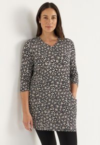 Lange Tunika aus dunklem Stoff mit einem Blumenmuster in Creme, Rosa und Grün. Sie verfügt über einen V-Ausschnitt und halblange Ärmel sowie seitliche Taschen.