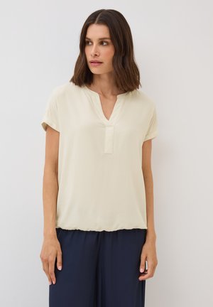 Light beige short-sleeve blouse with a V-neckline and seam detailing. Soft fabric with a relaxed fit, paired with navy pants.