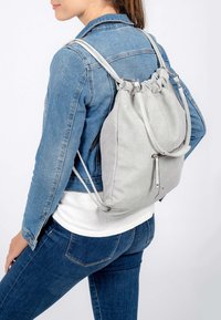 Sac à dos à cordon perforé gris avec des sangles lisses, maintenu par un matériel métallique, porté sur le dos d'une personne, contrastant avec une veste en denim.