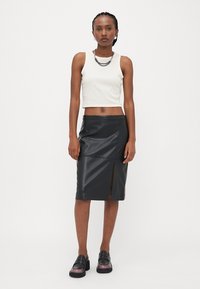 Svart fusk-läder blyertskjol med en framdelning, tillsammans med en ärmlös vit crop top. Modellen bär kraftiga svarta loafers.
