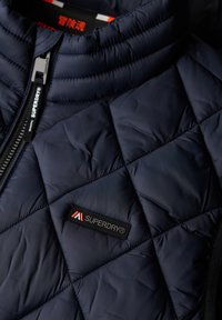 Marineblauwe gewatteerde jas met een hoge kraag, voorzien van een ritssluiting en een zwart logo-patch met het label "SUPERDRY" aan de voorkant.