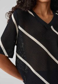 Top in rete nera con scollatura a V, caratterizzato da strisce diagonali bianche. Maniche corte, motivo a tessitura aperta e bordo nero a contrasto attorno al collo.