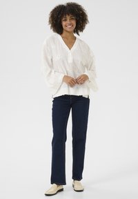 Blusa bianca ricamata con scollo a V e maniche a campana, abbinata a jeans dritti blu scuro e mocassini beige.