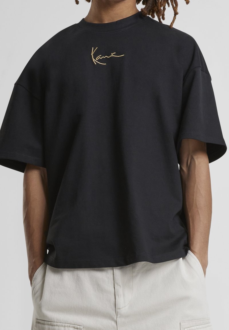 Persona wear un t-shirt negro de gran tamaño con una firma bordada en beige y pantalones blanco roto, con las manos en los bolsillos, en un fondo neutro.