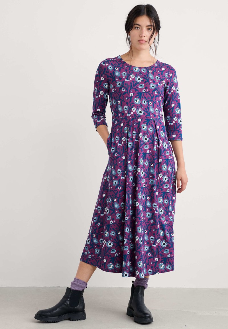 Paarse bloemenjurk met driekwartmouwen en een getailleerd lijfje, voorzien van een uitlopende rok. Gecombineerd met zwarte enkellaarsjes en paarse sokken.