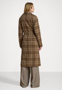 Cappotto lungo a quadri marrone in misto lana, con cintura, colletto a punte e tessuto testurizzato. Abbinato a pantaloni larghi color marrone chiaro.