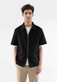 Svart kortärmad zip-up skjorta med subtil textur, bred krage och silverfärgad dragkedjedetalj, bärs över en vit T-shirt.