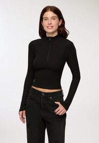 Młoda kobieta z długimi brązowymi włosami, ubrana w dopasowaną czarną bluzkę zapinaną na zamek z długim rękawem oraz czarne jeansy o wysokim stanie, pozująca z jedną ręką w kieszeni.