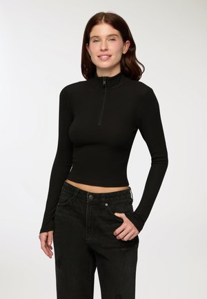 Giovane donna con lunghi capelli castani, che indossa una maglietta nera attillata con maniche lunghe e zip, e jeans neri a vita alta, in posa con una mano in tasca.