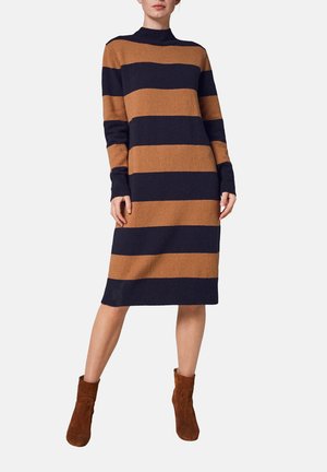 Kobieta ubrana w sukienkę swetrową o długości do kolan z szerokimi poziomymi granatowymi i brązowymi paskami, w komplecie z brązowymi botkami za kostkę.