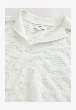 Cardigan en tricot blanc présentant un motif géométrique texturé, un col polo et trois boutonnières à l'avant.