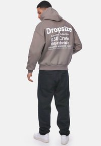 Grauer Kapuzenpullover mit weißem Text auf dem Rücken, mit abfallenden Schultern und gerippten Bündchen, kombiniert mit schwarzen Hosen und weißen Sneakers.