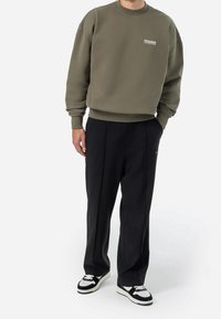 Sudadera verde oliva con puños y dobladillo acanalados, que presenta un logo blanco. Combinada con pantalones negros plisados y zapatillas blancas y negras.