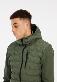 Veste doudoune verte olive avec capuche doublée, dotée d'une poche poitrine zippée et d'une texture matelassée. Tissu lisse sur les manches.