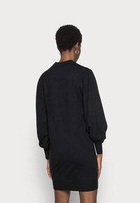 Personne avec des cheveux courts et bouclés portant une robe en maille noire à manches longues, vue de dos sur un fond clair uni.