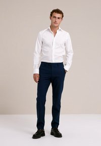 Balti, ilgomis rankovėmis mygtukinė palaidinė derinama su tamsiai mėlynomis kelnėmis. Palaidinėje yra klasikinis apykaklė, o kelnės pasiūtos pagal figūrą.