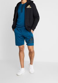 Conjunto deportivo masculino que incluye una sudadera con cremallera negra, una camiseta azul y pantalones cortos azules a juego con un logo negro, combinados con zapatillas blancas.