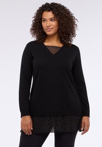 Czarna bluzka z długim rękawem i dekoltem w kształcie litery V, nałożona na przezroczystą, czarną warstwę z kropkami. Gładka faktura i luźny krój.